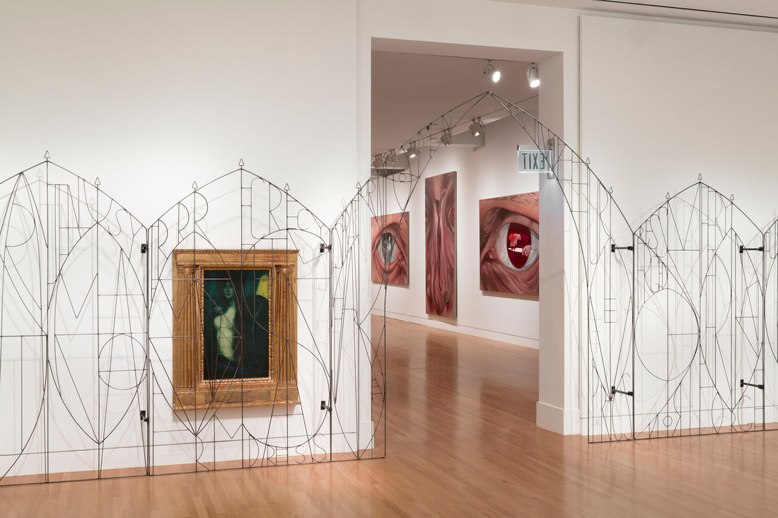 Image is a photo of a slim steel gate with letters across its form. Behind the gate on the wall is a painting of a woman, and a large doorway into another gallery revealing a glimpse of paintings of eyes and a nose.